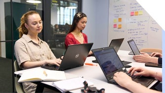 Two women on laptops during a team meeting with sticky notes in the background