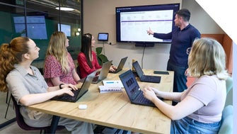 A group of professionals attending a presentation led by a confident presenter pointing at a screen displaying graphs.