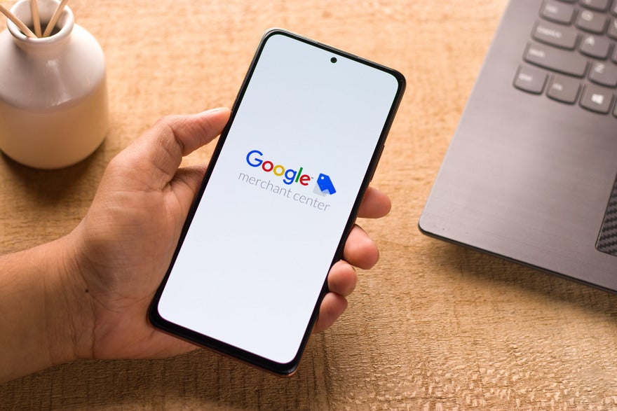 Hand holding a mobile with Google merchant center logo displayed on a white screen