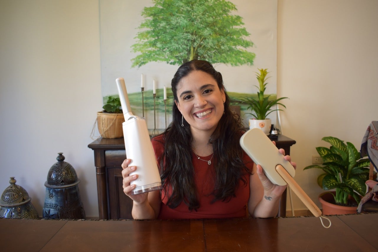 waist high headshot of woman holding two lint brushes smiling in front of wood table with tree painting behind