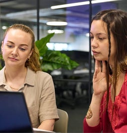 Two professional women collaborating on a project using a laptop in an office setting.