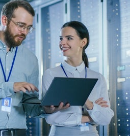 Two colleagues stand in data center holding laptop and smiling