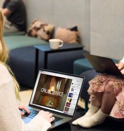 Two women using laptops in a modern office lounge area - one is watching an online service on YouTube.