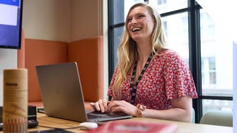 A joyful professional woman smiling while working on her laptop at an office desk.