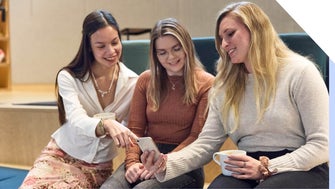 Three women smiling and looking at a smartphone together, with one woman pointing at something on the screen.