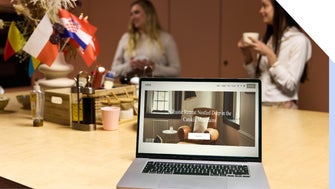 People chat in a kitchen setting with international flags, with a laptop in the foreground showing a rustic retreat in the Catskill Mountains.