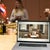 People chat in a kitchen setting with international flags, with a laptop in the foreground showing a rustic retreat in the Catskill Mountains.