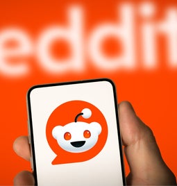 Hand holding a mobile showing a white background with the Reddit logo on, held against the red Reddit background