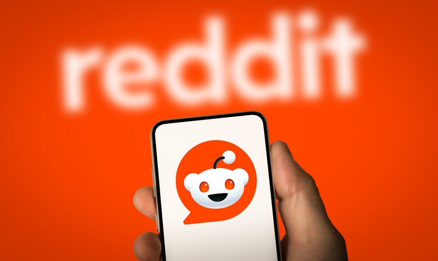 Hand holding a mobile showing a white background with the Reddit logo on, held against the red Reddit background