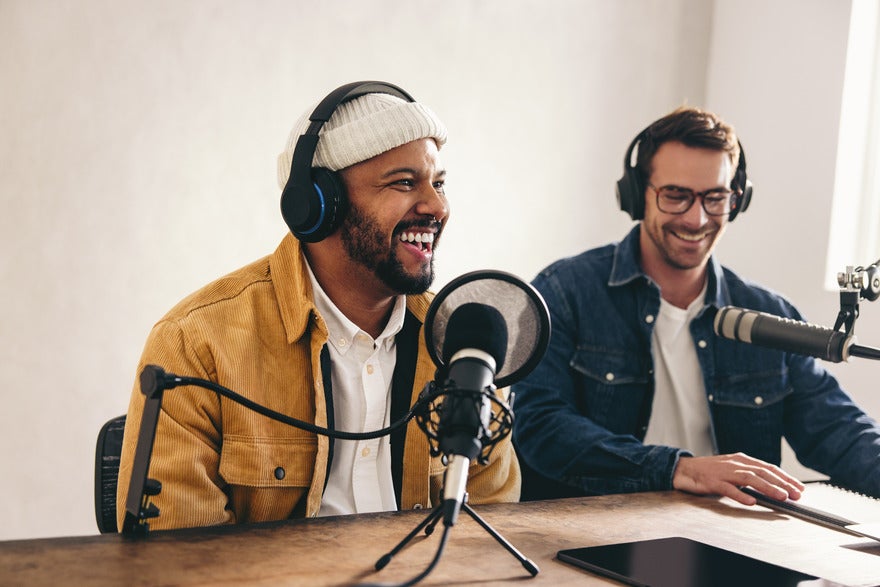 Two men sat a desk with podcast equipment in front of them and headphones on