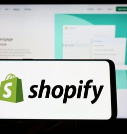 A phone with the shopify logo help in front of a blurred computer screen