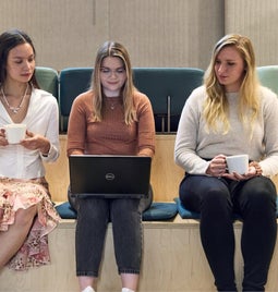 Three professional women engaged in work, with one using a laptop and two others discussing over coffee in a modern office lounge area.